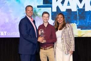 A man in a suit, on the left, hands an award to a man in a red shirt, center, and a women in a plaid jacket, on the right